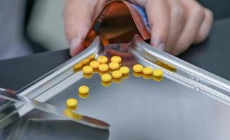 Selective focus on yellow tablets pills on stainless tray with blur hand of pharmacist or pharmacy technician counting pills into a plastic zipper bag. Prescription medicine. Medical health care. photo