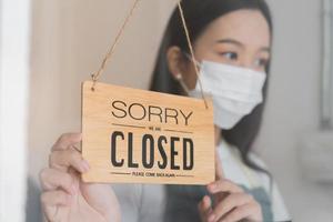 Asian young owner retail,coffee shop woman turning sign board to closed with wearing face mask,protection to pandemic of coronavirus.Close store,restaurant or pub due to lockdown, quarantine of covid. photo