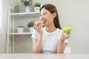 dieta, dieta asiático joven mujer o niña sonrisa, confuso elegir, elección verde manzana o rosado dulce rosquilla, a hogar, comer comida para bueno saludable, salud cuando hambriento. cerca arriba hembra peso pérdida persona. foto