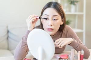 Beauty blogger concept, cute asian young woman, girl make up face by applying mascara brush on eyebrow, looking at the mirror at home. Female look with natural fashion style. photo