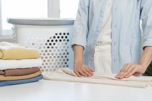 ama de casa, asiático joven mujer mano en muchos plegable recién camisas, suéteres o vestir en escritorio, mesa después Lavado limpiar ropa y el secado, haciendo casa trabajando en habitación a hogar. lavandería y mucama. foto