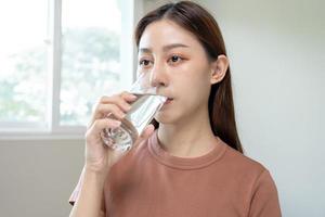 contento hermoso, asiático joven mujer, niña Bebiendo, sorbo Fresco vaso de agua para hidratación de cuerpo, participación transparente vaso en su mano, sediento a hogar. salud cuidado, sano estilo de vida concepto. foto