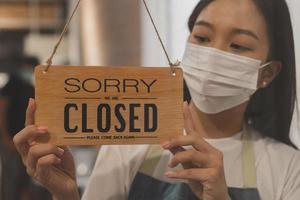 Asian young owner retail,coffee shop woman turning sign board to closed with wearing face mask,protection to pandemic of coronavirus.Close store,restaurant or pub due to lockdown, quarantine of covid. photo