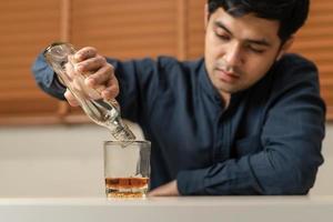 Alcoholism, depressed asian young man pouring, holding bottle whiskey into the glass, drinking alcohol  beverage at night. Treatment of alcoholic addiction, suffer abuse problem alcoholism concept. photo