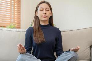 hermosa asiático joven mujer, niña manos en calma actitud sentado práctica meditando en loto posición en sofá a hogar, meditación, ejercicio para bienestar, sano cuidado. relajación, contento ocio gente. foto