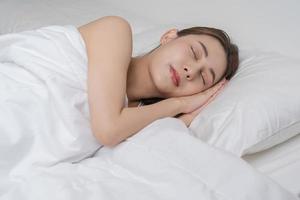 dulce Sueños, atractivo sonrisa bonito asiático joven mujer, niña dormido en cómodo cama acostado en suave almohada descansando, acuerdo ojos cerrado mientras con cubierto cobija en acogedor blanco cama en el Mañana. foto