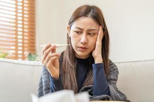 Sick, influenza asian young woman, girl headache have a fever, flu and check thermometer measure body temperature, feel illness sitting on sofa bed at home. Health care person on virus, covid-19. photo