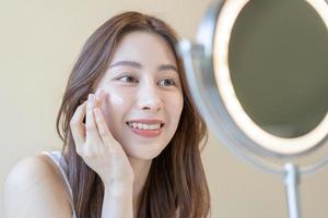 Fresh healthy skin, beautiful smile of asian young woman, girl looking at mirror, applying moisturizer on her face, putting cream treatment before make up cosmetic routine at home. Facial Beauty. photo