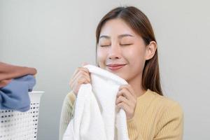 sensación blandura, sonriente asiático joven mujer, niña conmovedor mullido toalla algodón, oliendo Fresco limpiar ropa en mesa después Lavado, lavadero, seco. casa trabajando a hogar. lavandería y mucama. foto