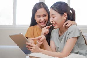 Happy lesbian, pleasure asian young two women, girl gay or close friend, couple love embrace, spending good time together, using tablet for entertainment on sofa at home. Activity of leisure, relax. photo