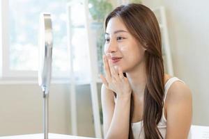 Fresh healthy skin, beautiful smile of asian young woman, girl looking at mirror, applying moisturizer on her face, putting cream treatment before make up cosmetic routine at home. Facial Beauty. photo