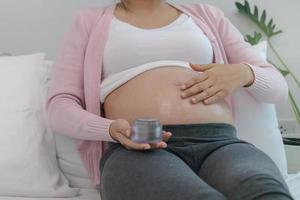 Motherhood asian young pregnant woman applying pregnancy lotion prevent stretch marks on belly by rubbing cream with moisturizer, moisture for abdomen, belly beauty skin, health care at home. photo