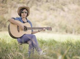 Women short hair wear hat and sunglasses sit playing guitar in grass field photo