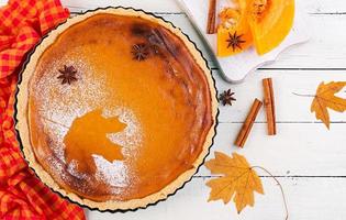 American homemade pumpkin pie with cinnamon and nutmeg, pumpkin seeds and autumn leaves on a white wooden table. Thanksgiving food. Top view. Flat lay photo