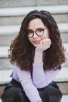 Beautiful young woman with brunette curly hair, portrait in eye glasses enjoying the sun in the city. photo