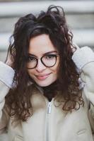 Beautiful young woman with brunette curly hair, portrait in eye glasses enjoying the sun in the city. photo