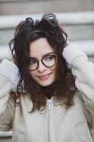 Beautiful young woman with brunette curly hair, portrait in eye glasses enjoying the sun in the city. photo