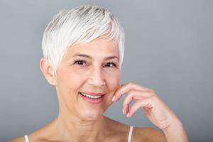 Senior woman pulling cheeks to feel softness and looking at camera. Beauty portrait of happy mature woman smiling with hands on cheek isolated over grey background. Aging process and skin concept. photo