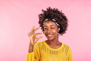 Size matters. Pleased African American young woman demonstrates very tiny object, smiles positively, wears casual sweater, poses against pink background, shapes small thing. Body language concept photo