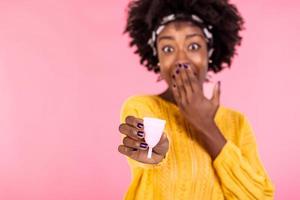 Serious dark skinned woman holds bell shaped menstruation cup for inserting in vagina, trapping menstrual fluid and leakage protection, tells secret information and tips how to use it, makes hush sign photo