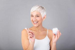 Beautiful mature woman holding jar of skin cream for face and body isolated on grey background. Happy senior woman applying anti-aging moisturizer and looking at camera. Beauty anti aging treatment. photo