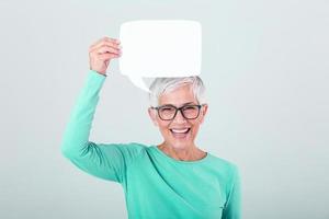 Happy mature woman in plain blue long sleeve t-shirt holding empty speech bubble isolated on background. Woman showing sign speech bubble banner looking happy and pointing her finger on it photo