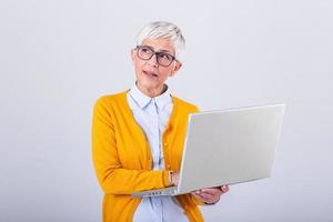 Photo of a thinking mature business woman isolated over grey background holding laptop computer. Image of confused senior woman using laptop computer. Looking at laptop while holding face with hands.