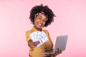 Image of cheerful African American woman standing isolated over pink background using laptop computer and holding money banknotes . Portrait of a smiling girl holding laptop computer photo