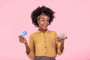 Portrait of a African American woman standing on a pink background holding smartphone and money, cashback. woman holding money and paying online on her mobile phone photo