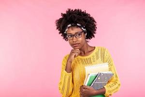 Beautiful young african american woman holding notebooks, wearing glasses. Having thinking face. Student life photo