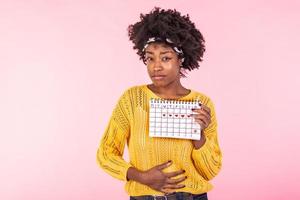 Young African American woman suffering from belly pain and showing her periods calendar isolated over pink background. Medical, healthcare, gynecological concept. Copy space photo