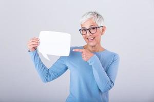 Happy mature woman in plain blue long sleeve t-shirt holding empty speech bubble isolated on background. Woman showing sign speech bubble banner looking happy and pointing her finger on it photo