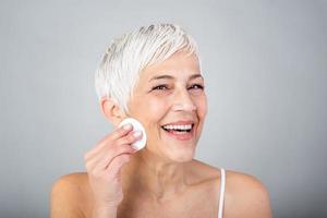 Healthy mature woman removing makeup from her face with cotton pad isolated on grey background. Beauty portrait of happy woman cleaning skin and looking at camera. photo