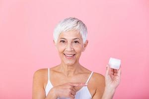 Beautiful mature woman holding jar of skin cream for face and body isolated on grey background. Happy senior woman applying anti-aging moisturizer and looking at camera. Beauty anti aging treatment. photo