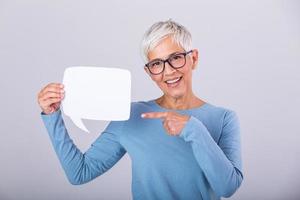 Happy mature woman in plain blue long sleeve t-shirt holding empty speech bubble isolated on background. Woman showing sign speech bubble banner looking happy and pointing her finger on it photo