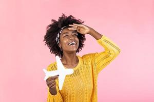 Photo of African American positive cute pretty beautiful girl won tickets to go abroad by air plane rejoicing excited emotions holding plane isolated curly wavy pink vivid color background