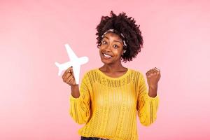 Photo of African American positive cute pretty beautiful girl won tickets to go abroad by air plane rejoicing excited emotions holding plane isolated curly wavy pink vivid color background
