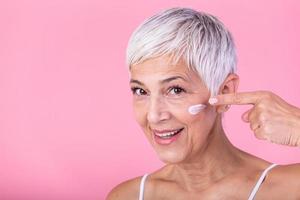 Beautiful mature woman holding jar of skin cream for face and body isolated on pink background. Happy senior woman applying anti-aging moisturizer and looking at camera. Beauty anti aging treatment. photo