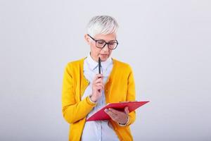 Photo of a thinking mature business woman isolated over grey background holding clipboard and pen. Image of confused senior woman looking at documents