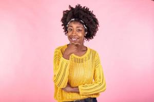 Adorable young black woman with natural beauty, pleasant smile, loooks happily, smiles gently, wears yellow poloneck, has curly bushy hair, isolated over pink background. Pleasant emotions concept