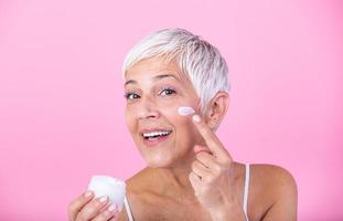 Beautiful mature woman holding jar of moisturizer and looking at camera. Happy senior woman holding a bottle of anti-aging lotion isolated over pink background. Anti aging and beauty treatment. photo