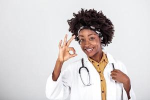 contento africano hembra médico vestir blanco laboratorio abrigo, estetoscopio Mira a cámara y demostración Okay signo. sonriente afro americano mujer profesional terapeuta médico retrato. foto