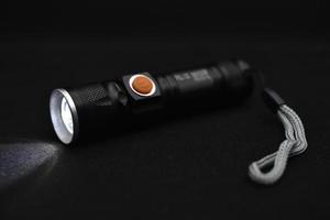 Metal black flashlight on a black background. Burning pocket flashlight close-up. Lighting device. photo