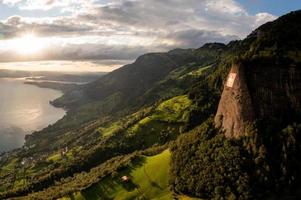 a huge swiss flag is fixed on a cliff, below it is a village on a lake photo