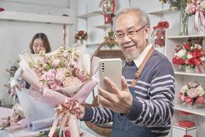 Happy Asian senior male florist owner selfie pictures and show floral bouquet arrangement with smartphone in valentine at bright flower shop, beautiful blossoms store, small business SME entrepreneur. photo