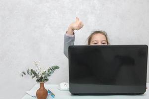 little girl sits at a table peeking out from behind a laptop and shows like, online learning photo