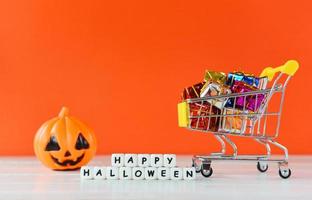 Halloween shopping holiday concept - word blocks happy halloween decorations and pumpkin jack o lantern with gift box in a shopping cart on orange background