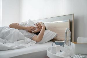 asiático mujer tos, estornudos y conmovedor frente mientras sensación enfermo y acostado en cama con termómetro y pastillas en mesa foto