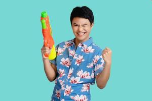 Happy Asian young man holding a water gun isolated on bright blue background. He is smiling and shows signs yes by hand. photo