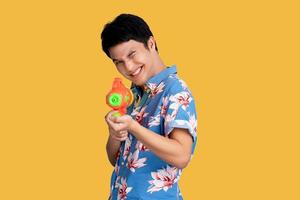 Young Asian man wearing colorful shirt having fun using a water gun on a yellow background. photo